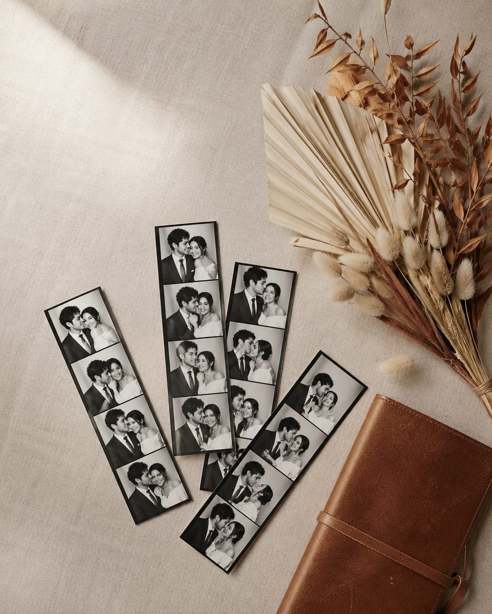 Editorial flat-lay of black-and-white wedding photobooth strips on bone linen with cognac leather notebook and dried palm botanicals, Soft Hours.